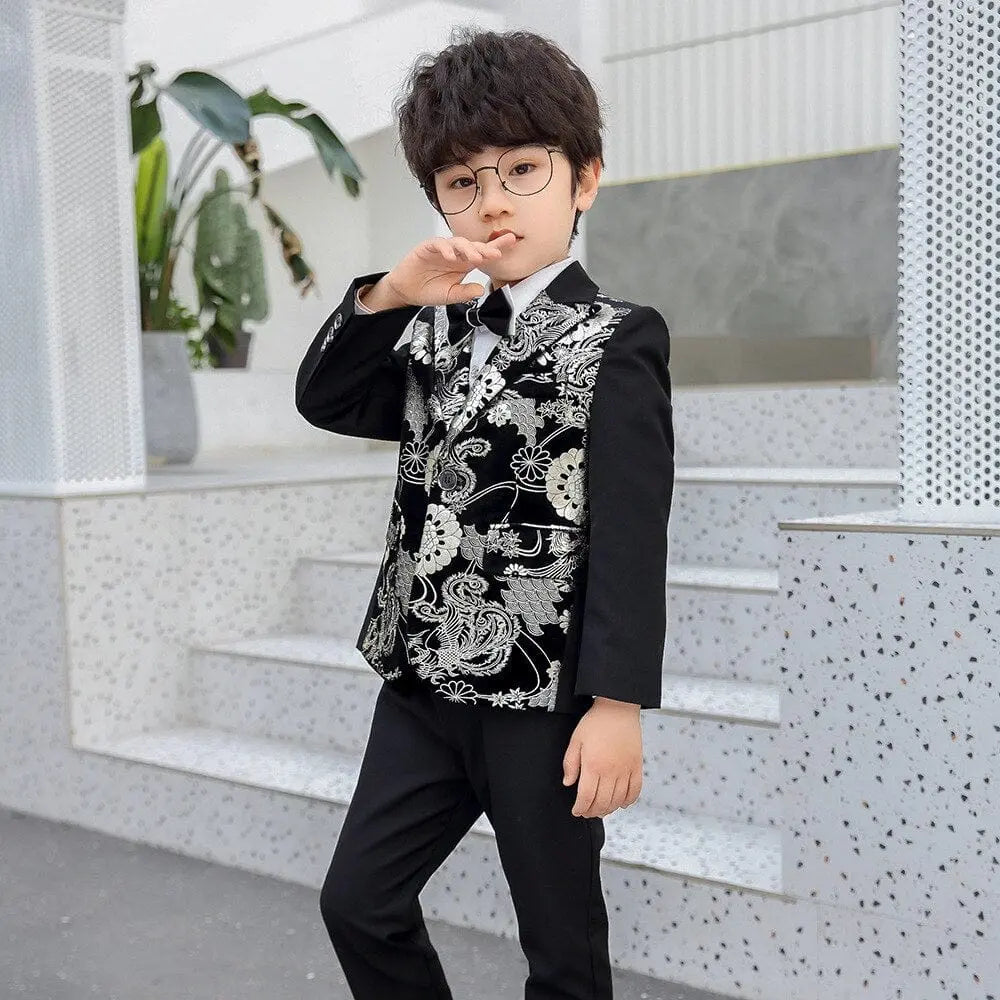 A young boy with glasses poses on steps, wearing the Children Formal Dress Kids Flower Jacket Pants Bowtie 3PCS Piano Costume—a bronzing show host photography suit. White and gray terrazzo surfaces and potted plants enhance the stylish background.