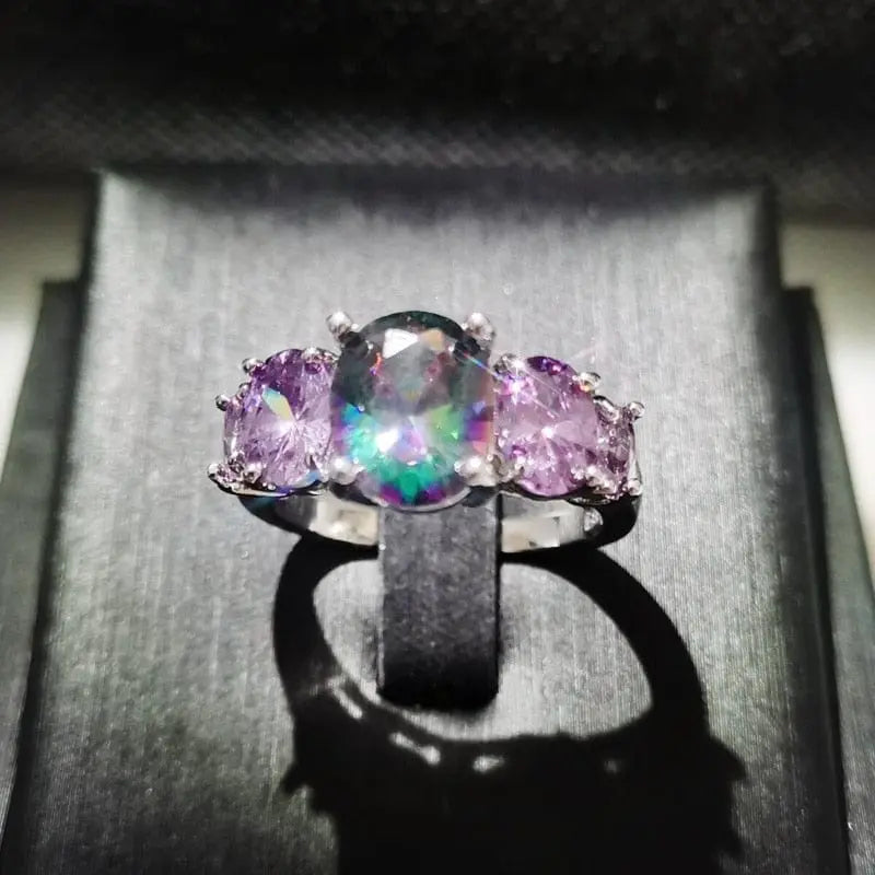 The CiNily Luxurious Mystery Stone Ring features a silver-plated band with a large iridescent rainbow stone and two smaller oval purple stones, beautifully displayed on a black stand under a soft spotlight.