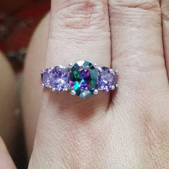 A hand displays the CiNily Luxurious Mystery Stone Ring—silver plated, featuring a large oval rainbow stone flanked by purple gems. Perfect as a fashion jewelry gift for women.