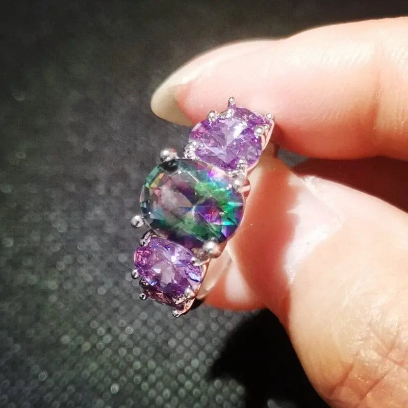 A hand holds the CiNily Luxurious Mystery Stone Ring, a silver-plated fashion ring with a central oval rainbow topaz and two square-cut purple stones—perfect for jewelry lovers.