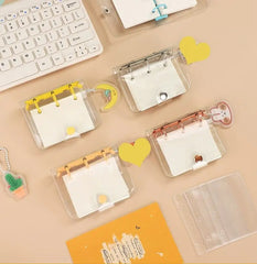 Several transparent mini three-ring binders with decorative charms are arranged on a desk next to a keyboard.