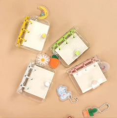 Four small, transparent three-ring mini notebooks with colorful binders and decorative charms.