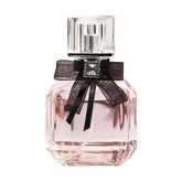 A clear glass perfume bottle filled with pink liquid, featuring a black bow and a square cap.