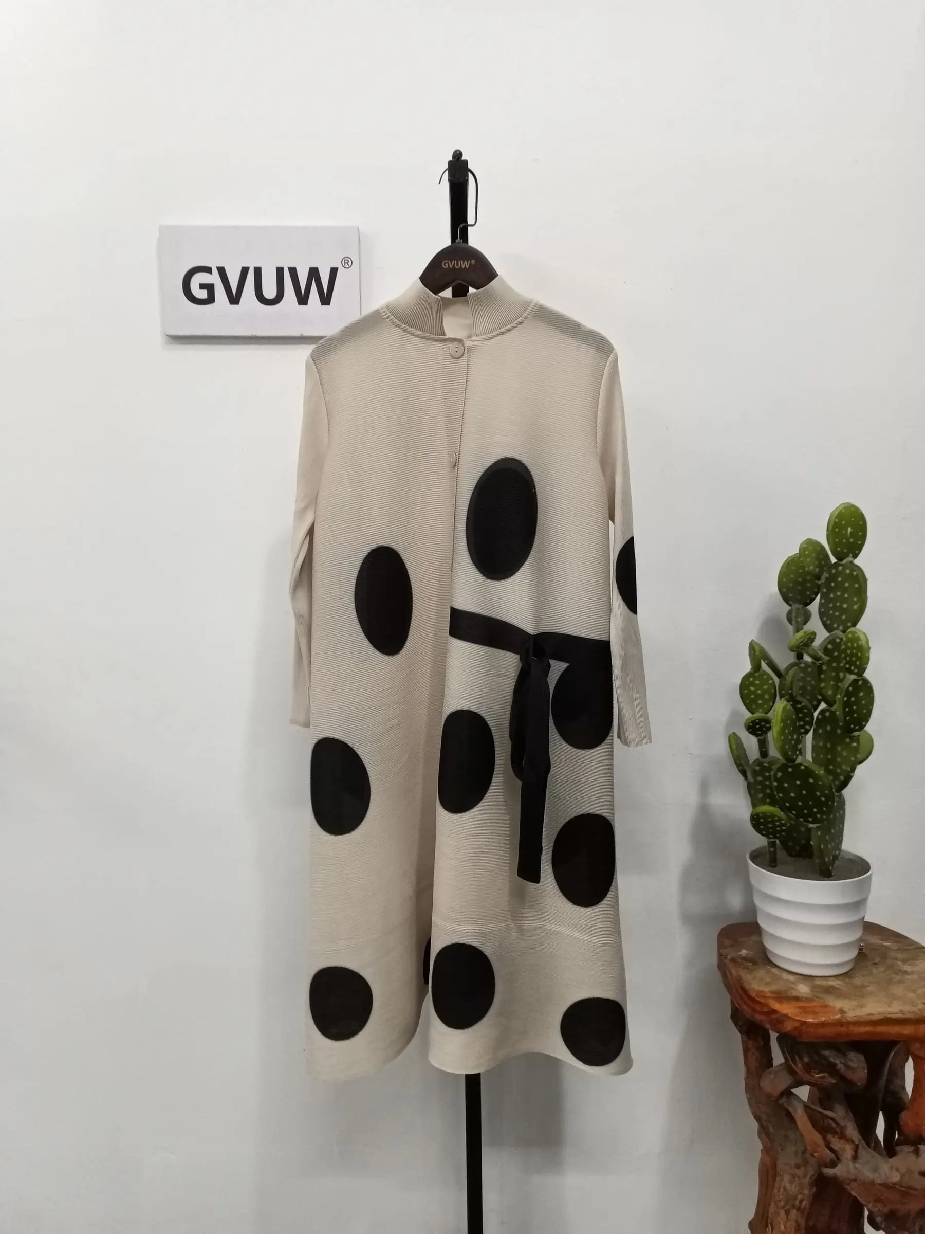 The Elegant Stand Collar Pleated Dress, featuring cream fabric with bold black polka dots, long sleeves, and a black belt, hangs on display. A potted cactus sits on a nearby stool beneath a GVUW sign.
