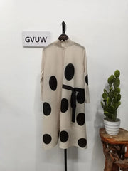 The Elegant Stand Collar Pleated Dress, featuring cream fabric with bold black polka dots, long sleeves, and a black belt, hangs on display. A potted cactus sits on a nearby stool beneath a GVUW sign.
