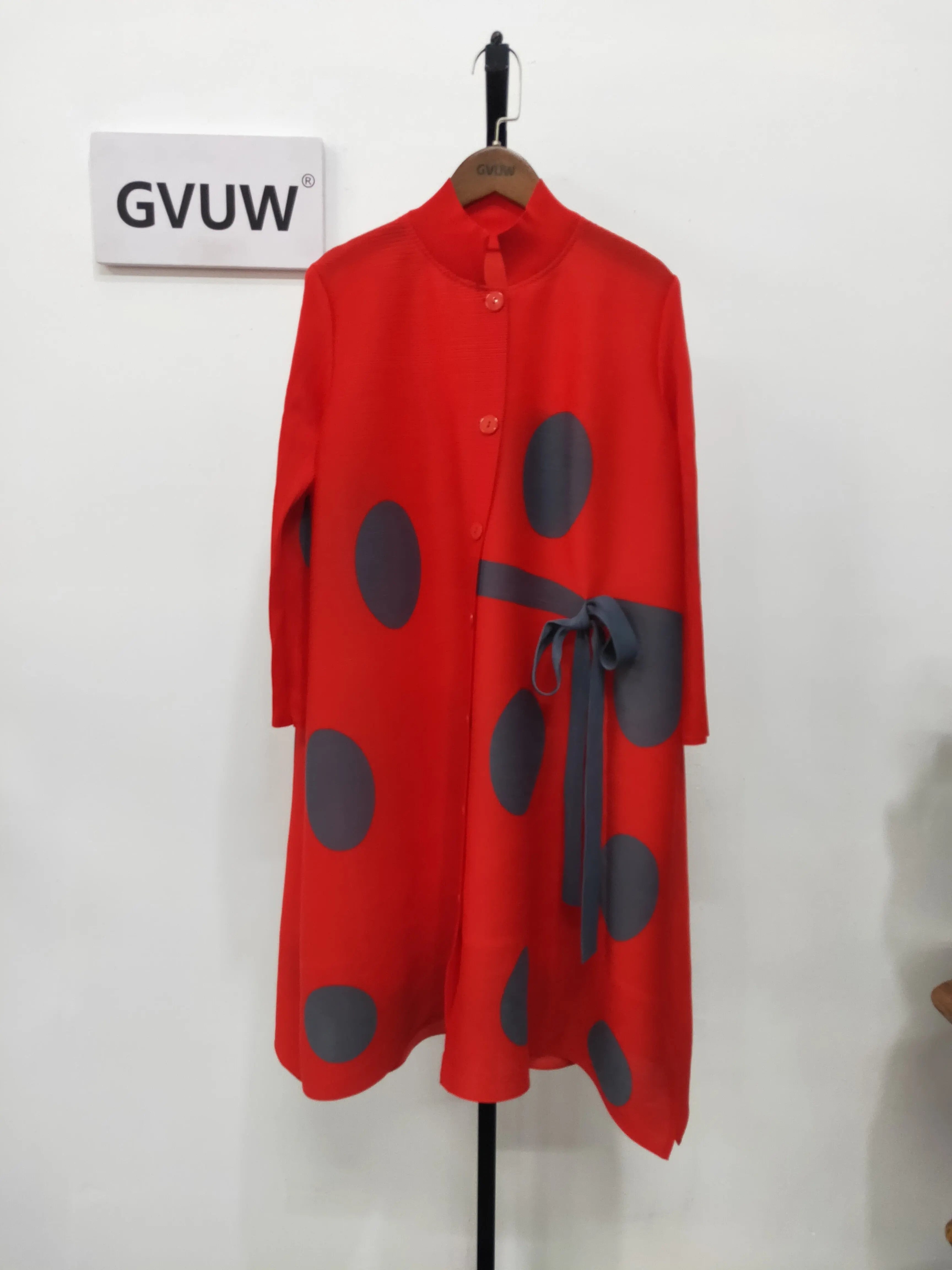 An Elegant Stand Collar Pleated Dress with red long sleeves, large black polka dots, and a tied sash hangs on a wooden hanger against a white wall beside a sign reading GVUW.