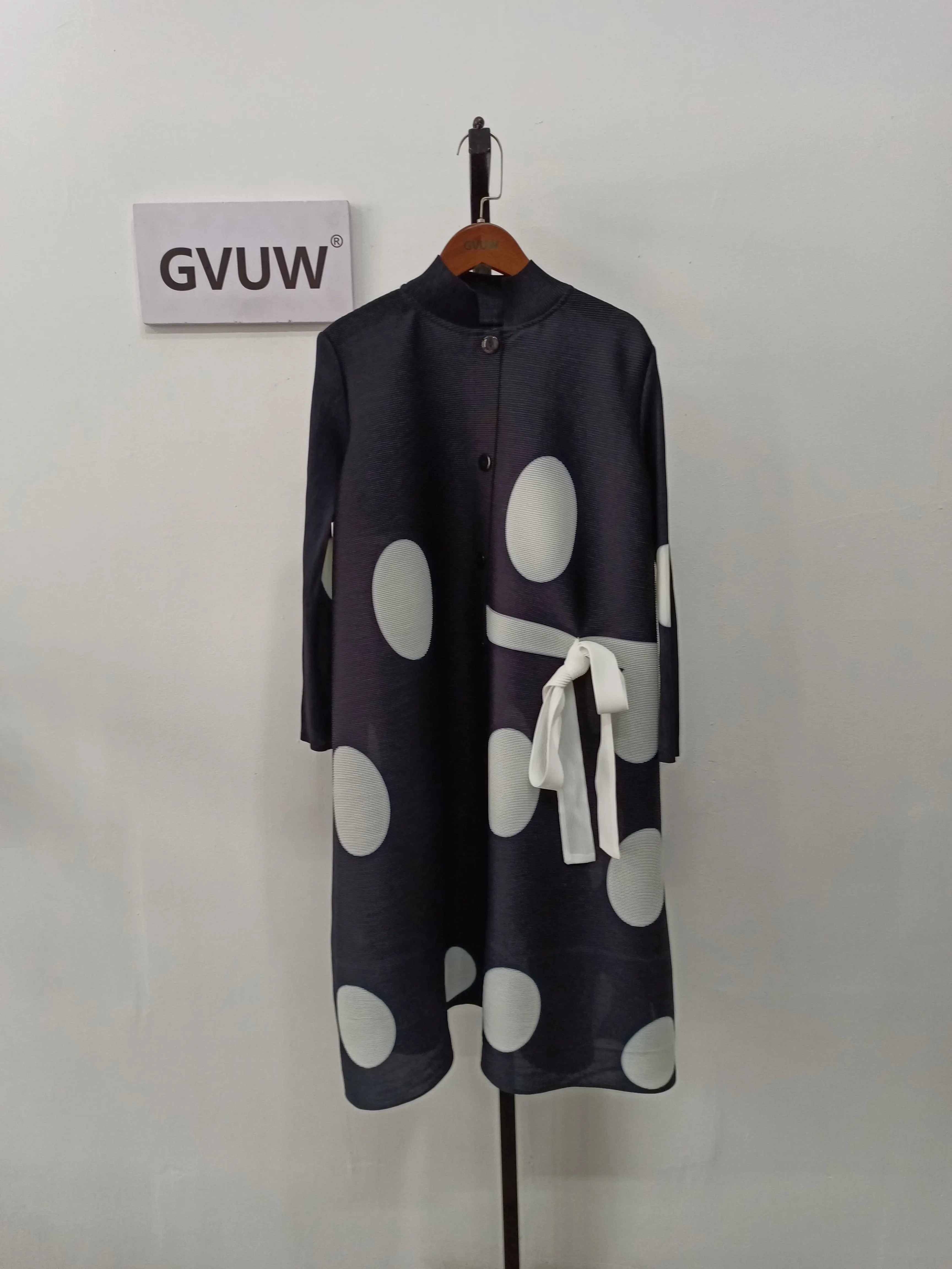 An Elegant Stand Collar Pleated Dress in black with large white polka dots, a white waist bow, and knee-length cut is displayed on a hanger. A sign reading GVUW is mounted on the wall behind it.