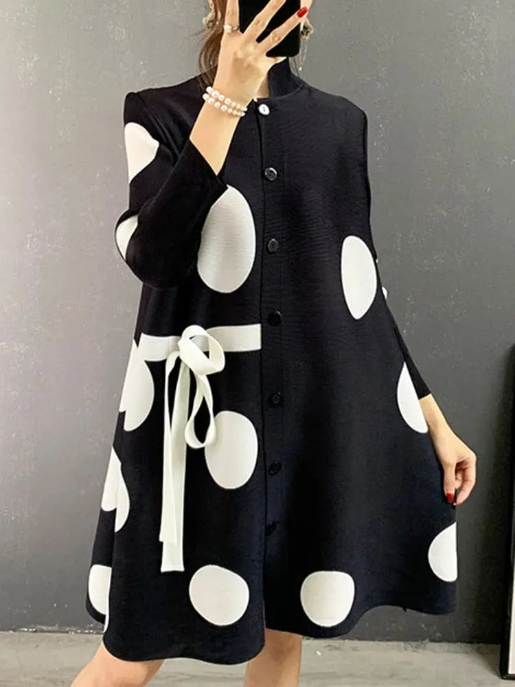 A person wears the Elegant Stand Collar Pleated Dress featuring a loose fit, dot print, front buttons, white waist bow, and long sleeves while standing against a plain gray wall.