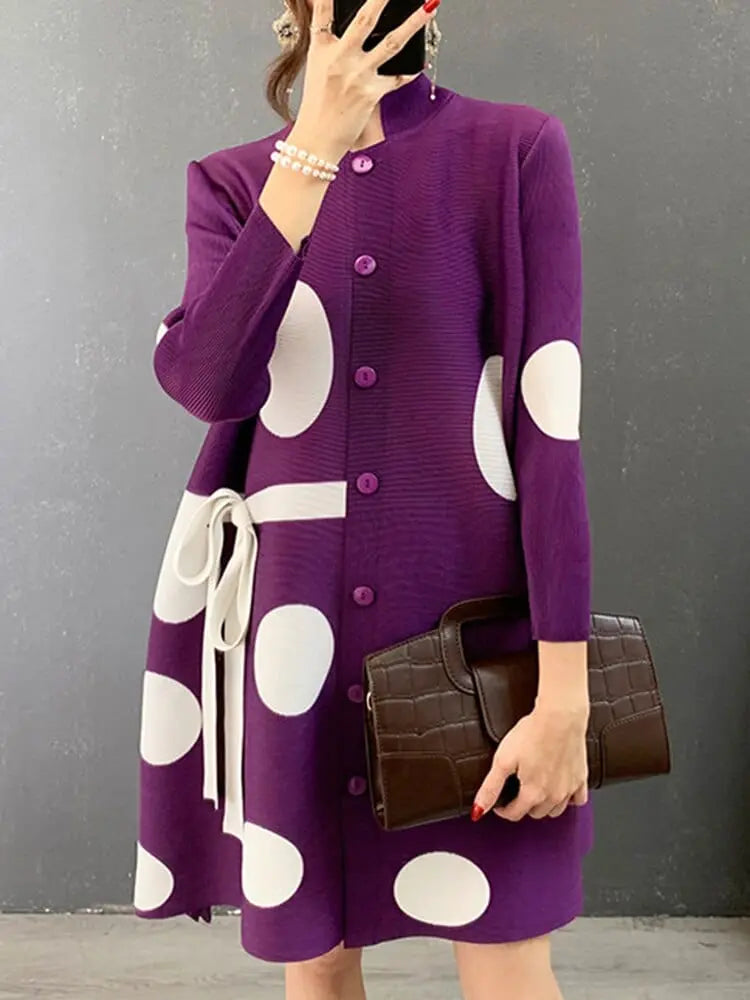 A person in the Elegant Stand Collar Pleated Dress with matching buttons and a white belt stands against a grey wall, holding a dark brown textured handbag.