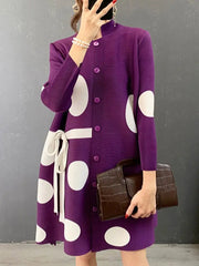 A person in the Elegant Stand Collar Pleated Dress with matching buttons and a white belt stands against a grey wall, holding a dark brown textured handbag.