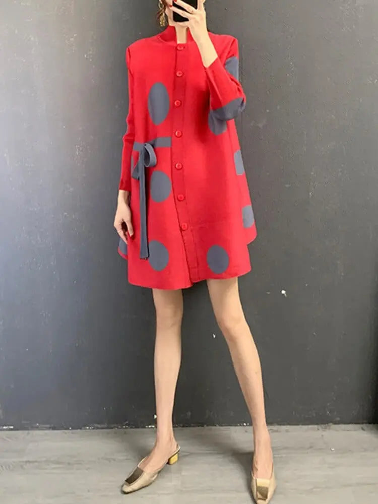 A person stands against a gray wall wearing the Elegant Stand Collar Pleated Dress, which is red with large gray polka dots, a matching belt, and knee-length long sleeves. Beige low-heeled shoes complete the look. The person's face is not visible.