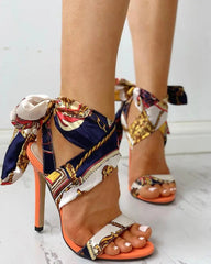 A pair of orange stiletto sandals featuring a patterned silk scarf tie around the ankle.