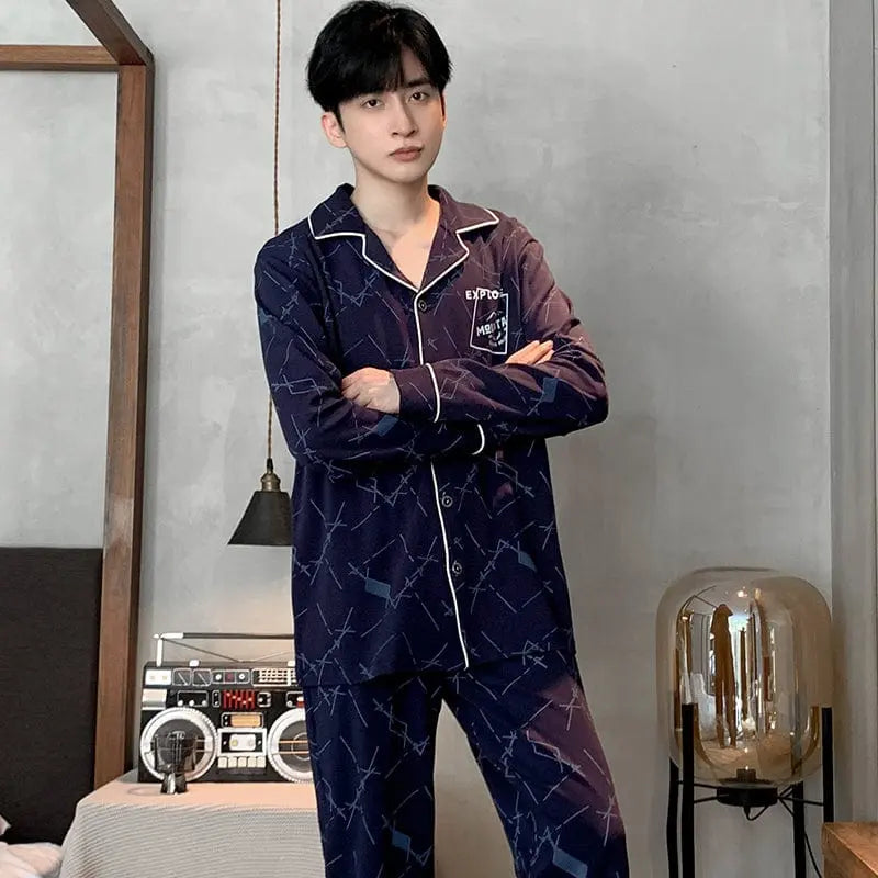 A young man with short black hair stands indoors wearing Factory wholesale winter 100% cotton men's pajamas—lapel cardigan, long sleeves, casual plus size sleepwear in dark blue geometric print—beside a table with a retro radio and glass lamp; gray wall and bed in background.