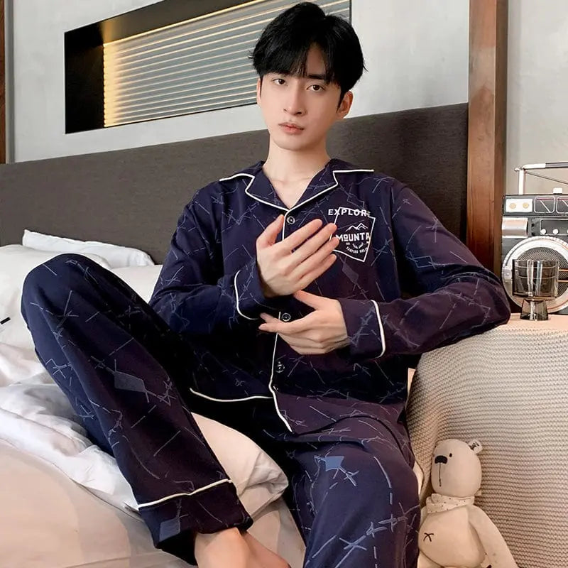 A young person with short black hair sits on a bed wearing Factory wholesale winter 100% cotton men's pajamas—a dark blue, geometric-patterned, lapel cardigan long sleeve nightwear. A small stuffed bear rests beside them.