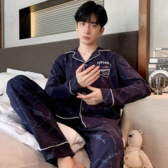 A young person with short black hair sits on a bed wearing Factory wholesale winter 100% cotton men's pajamas—a dark blue, geometric-patterned, lapel cardigan long sleeve nightwear. A small stuffed bear rests beside them.