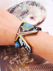 A close-up shows a wrist adorned with the Fashion Leopard Leather Bracelet for Women, featuring a wide leather band and metallic V-shaped charm. The blurred background includes magazine pages and a face partly hidden by flowers.
