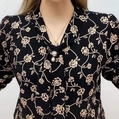 A black V-neck blouse with a gold floral print, featuring a pearl-adorned tie at the neckline.