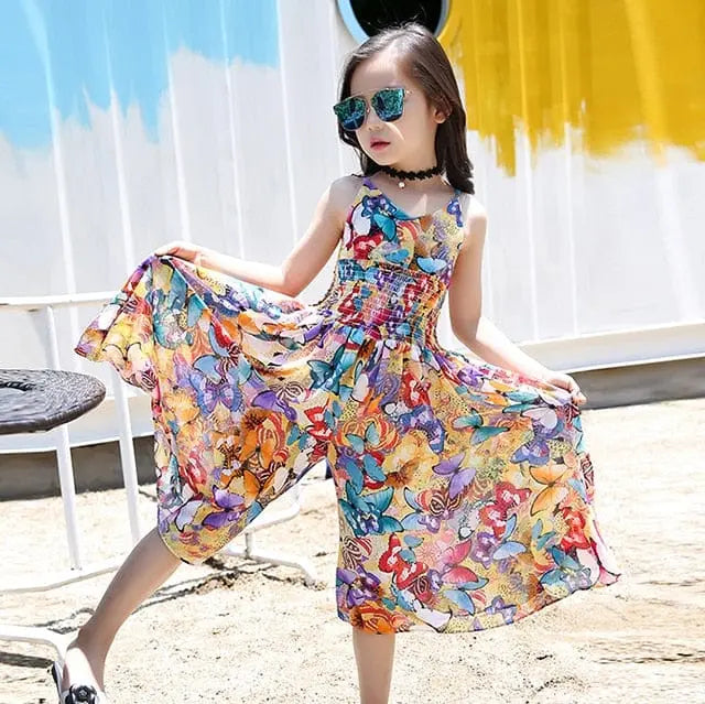 A girl wearing the Girls Flower Dress Kids Girl Beach Dresses Chiffon poses outdoors, holding the sides of her sleeveless floral dress beside a table, with a white and yellow painted wall in the background.