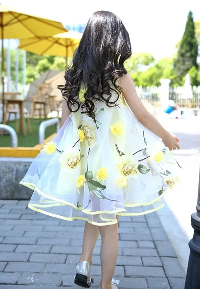 A young girl with long, wavy dark hair wears a Girls Flower Dress Kids Girl Beach Dress with floral print. She is walking away on a stone path outdoors near yellow umbrellas and green trees on a sunny day.