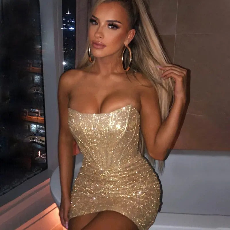 A woman with long blonde hair in a high ponytail wears the Giyu Night Club Party Sexy Bodycon Glitter Strapless Mini Dress, sitting on a bathtub edge in a modern bathroom at night and accessorizing with large hoop earrings.