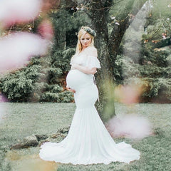 A pregnant woman wears a white off-shoulder mermaid maternity gown with ruffled sleeves.