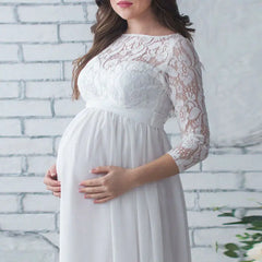 A pregnant woman wears a white ankle-length maternity dress with lace sleeves and a chiffon skirt.