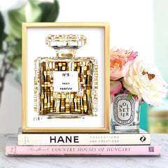 A framed gold-toned perfume bottle art print sits on a stack of books next to a floral candle.