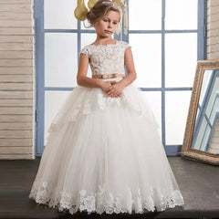 A girl wears a white floor-length ball gown with lace appliques and a tan sash at the waist.