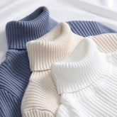 Three ribbed turtleneck sweaters in blue, beige, and white stacked on a white surface.