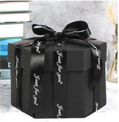 A black Hexagon Explosion Box Scrapbook Album with a lid is tied with a shiny black ribbon reading "Just for you" in white. The DIY handmade gift box rests on a marble surface, with stacked books in the background.