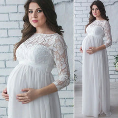 A pregnant woman wears a white ankle-length maternity dress with lace sleeves and a boat neckline.