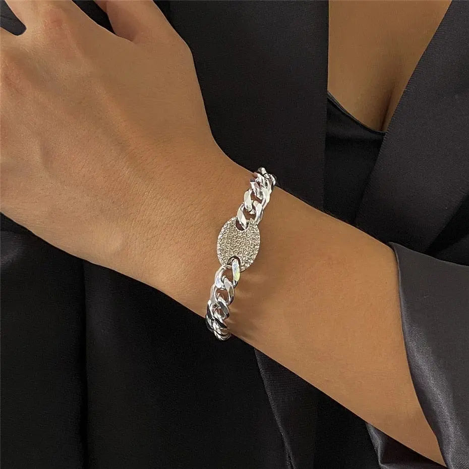 A person wears a silver chain bracelet with a round textured charm and the Ingemark Iced Out Rhinestones Tennis Chain Pendant Choker Necklace, styled against a dark satin outfit with a deep neckline.