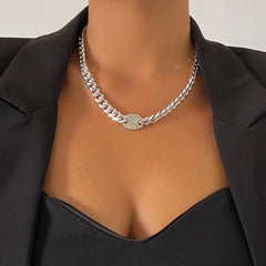 A person in a black blazer and strapless top showcases the Ingemark Iced Out Rhinestones Tennis Chain Pendant Choker—a chunky, silver Cuban CZ chain with a textured oval pendant, perfect for hip-hop or steampunk style.