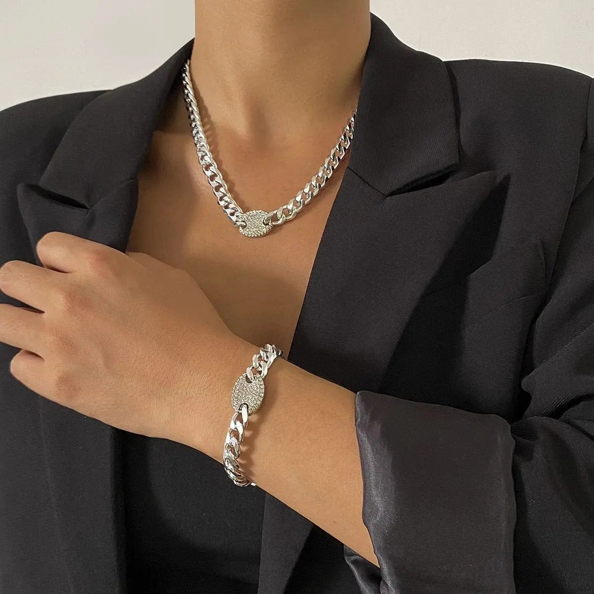A person in a black blazer showcases the Ingemark Iced Out Rhinestones Tennis Chain Pendant Choker Necklace, Cuban CZ Bling Jewelry, and matching bracelet with textured oval centerpieces against a plain light background.