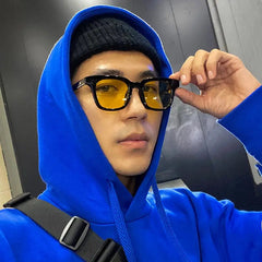 A person wearing square-framed sunglasses with yellow lenses and a black frame.