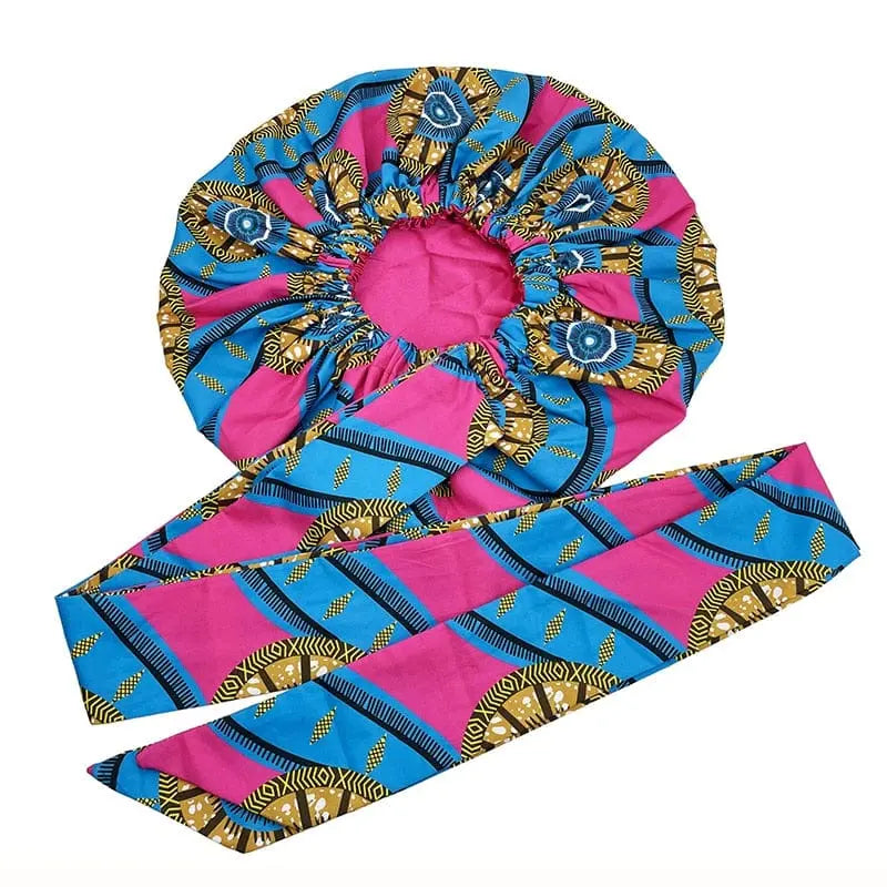 The New African Ankara Pattern Satin Lined Bonnet features a long ribbon headwrap and double-layer design, showcasing pink, blue, yellow, and black African geometric patterns. Displayed on a white background.
