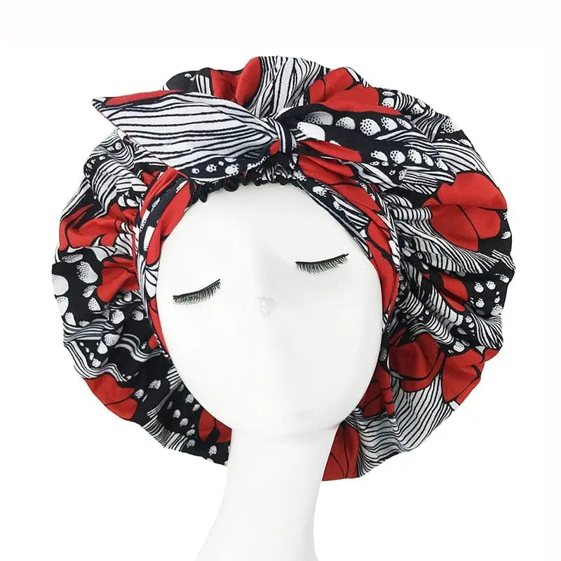 A mannequin head with closed eyes displays the New African Ankara Pattern Satin Lined Bonnet, a big-size adult hair cover featuring bold red and black patterns, stripes, double layer, long ribbon headwrap, and a prominent front bow.