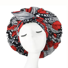 A mannequin head with closed eyes displays the New African Ankara Pattern Satin Lined Bonnet, a big-size adult hair cover featuring bold red and black patterns, stripes, double layer, long ribbon headwrap, and a prominent front bow.