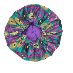 The New African Ankara Pattern Satin Lined Bonnet is a big-size women’s headwrap with a vibrant purple, green, yellow, and black pattern, soft purple inner lining, elastic opening, and long ribbon. Displayed on a white background.