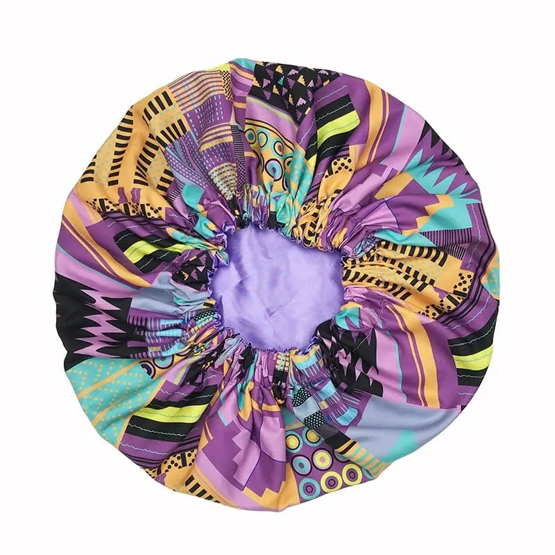 The New African Ankara Pattern Satin Lined Bonnet is a vibrant women’s headwrap with purple, yellow, green, and black geometric patterns, a gathered elastic opening, light purple inner lining, and long ribbon ties.