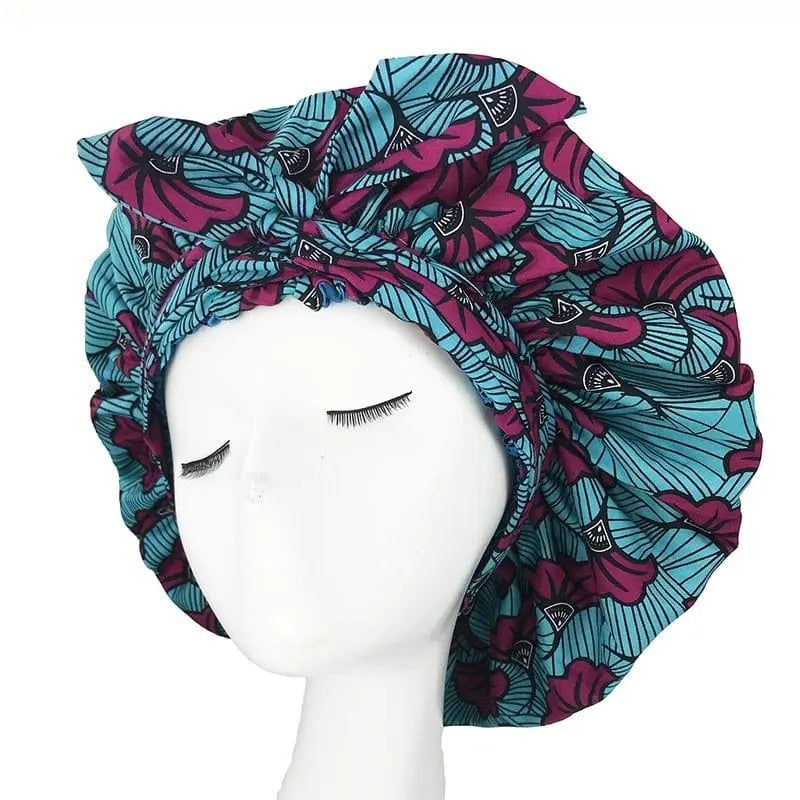 A mannequin head displays the New African Ankara Pattern Satin Lined Bonnet, a double-layer headscarf with a bold blue and purple floral pattern, long ribbon design, and big size for adult hair cover. The mannequin features closed eyes and long dark lashes.