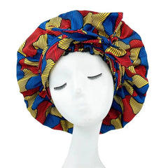 A mannequin head with closed eyes displays the New African Ankara Pattern Satin Lined Bonnet, a women’s big size double layer headscarf with bold red, yellow, and blue leaf patterns and a long ribbon.