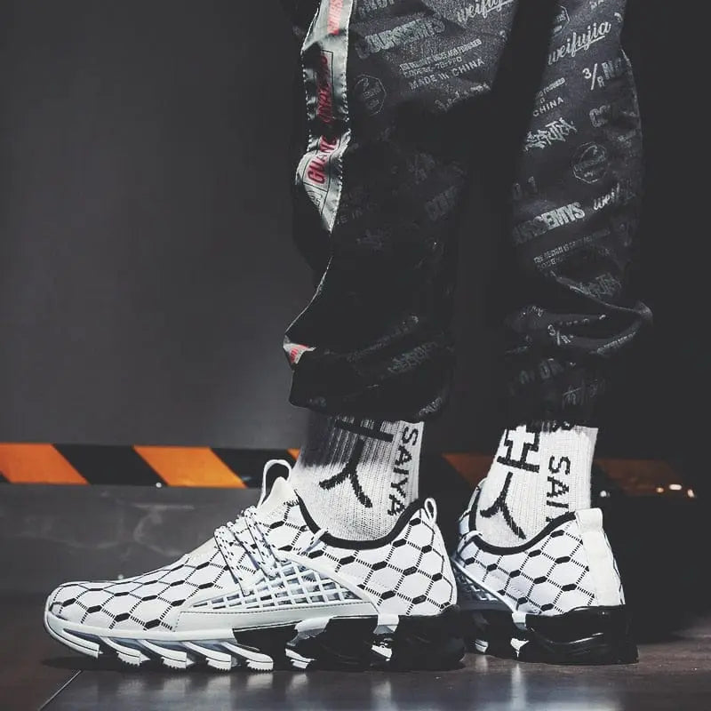 A person in black patterned joggers and white socks stands indoors near a yellow-black striped barrier, showcasing the New Fashion Men Running Shoes Sports Shoes Casual Trainers Mesh Tennis Sneakers Zapatos De Hombre.