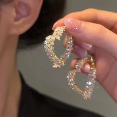 A hand holds a pair of gold-toned heart-shaped hoop earrings encrusted with sparkling crystals.