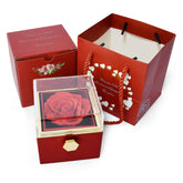 A red rose in a clear-topped jewelry box sits beside a matching gift box and a red paper bag.