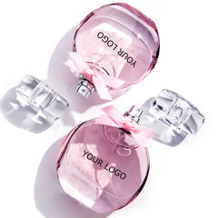 Two round, pink Perfume Customize Luxury Fragrance bottles with clear lids and pink bows are displayed on a white background. These long-lasting EDP luxury perfumes offer private label and OEM options for customized, fashionable scents.