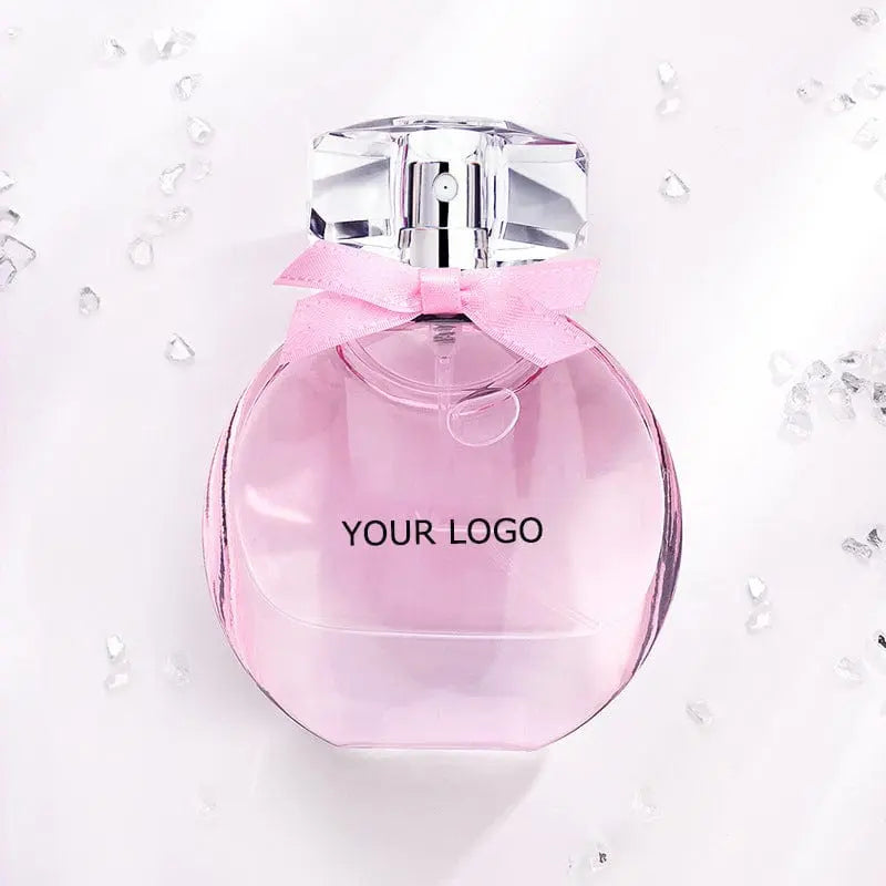 A round, pink Perfume Customize Luxury Fragrance bottle with a clear, faceted cap and light pink bow rests among small crystals on white, showcasing its delicate, feminine design. This long-lasting EDP is available for private label and OEM customization.