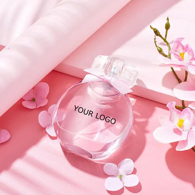 The Perfume Customize Luxury Fragrance sits on a pink surface amid pink and white flowers and soft fabric, with gentle sunlight casting shadows—ideal for scenes needing a touch of soft elegance.