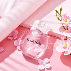 The Perfume Customize Luxury Fragrance sits on a pink surface amid pink and white flowers and soft fabric, with gentle sunlight casting shadows—ideal for scenes needing a touch of soft elegance.