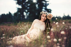 A pregnant woman wears a long-sleeved, ankle-length white lace maternity gown in a field.